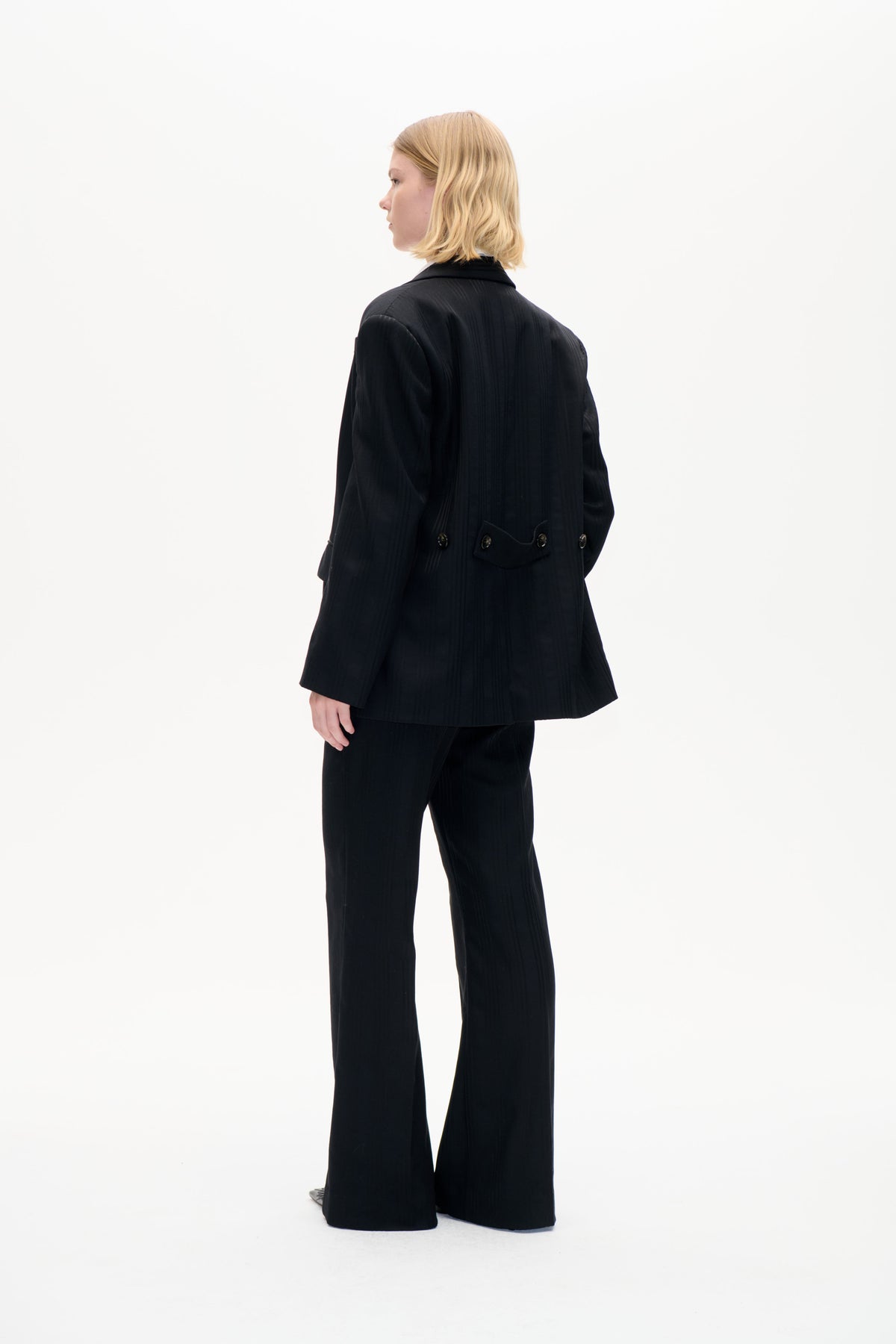Modell i svart kritstrecksrandig blazer med knäppt midjeband baktill och vida byxor, visad från sidan och bakifrån.