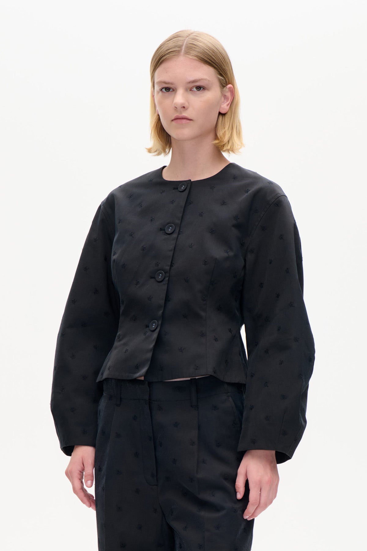Modellen bär en croppad svart jacka med subtila broderade motiv, rund halsringning och stora knappar, tillsammans med matchande byxor.