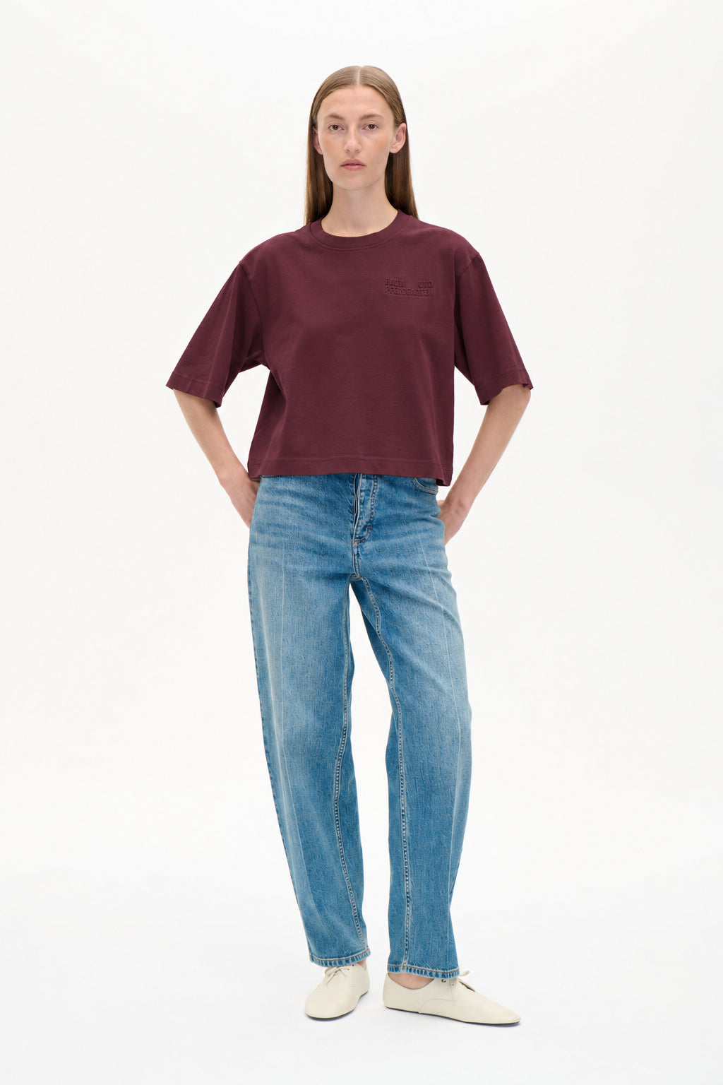 Framifrån på en modell som bär en vinröd kortärmad t-shirt med en ton-i-ton logotyp på bröstet och ljusblå mönstrade vida jeans.