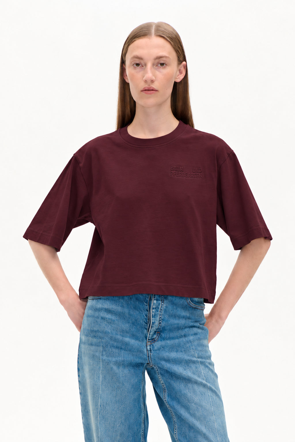 Modell i en croppad, kortärmad t-shirt i bordeaux med präglad logotyp på bröstet och ljusblå mönstrade jeans.