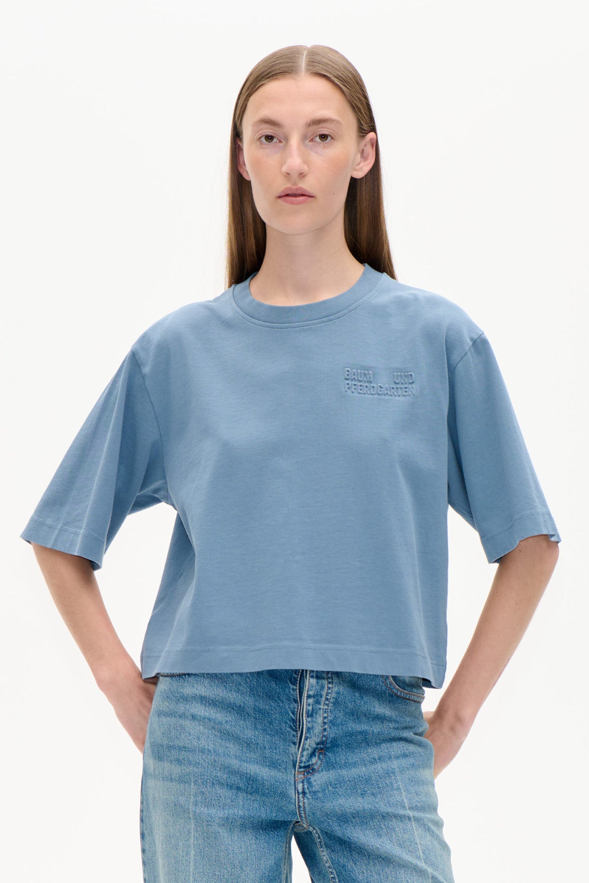 Framifrån: en modell i ljusblå oversized kortärmad t-shirt med upphöjd logga på bröstet och blå jeans.