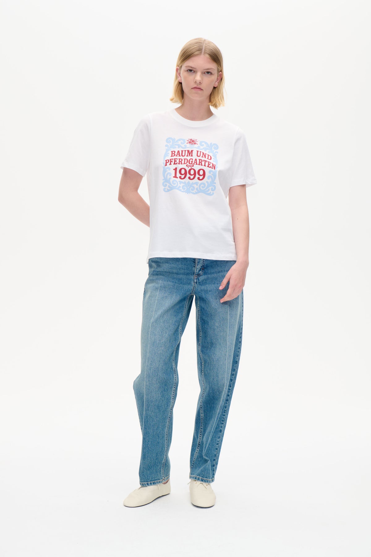 Modellen bär en vit kortärmad t-shirt med ett ornamentmotiv i blått och rött med texten 'Baum und Pferdgarten 1999', kombinerad med blå jeans.