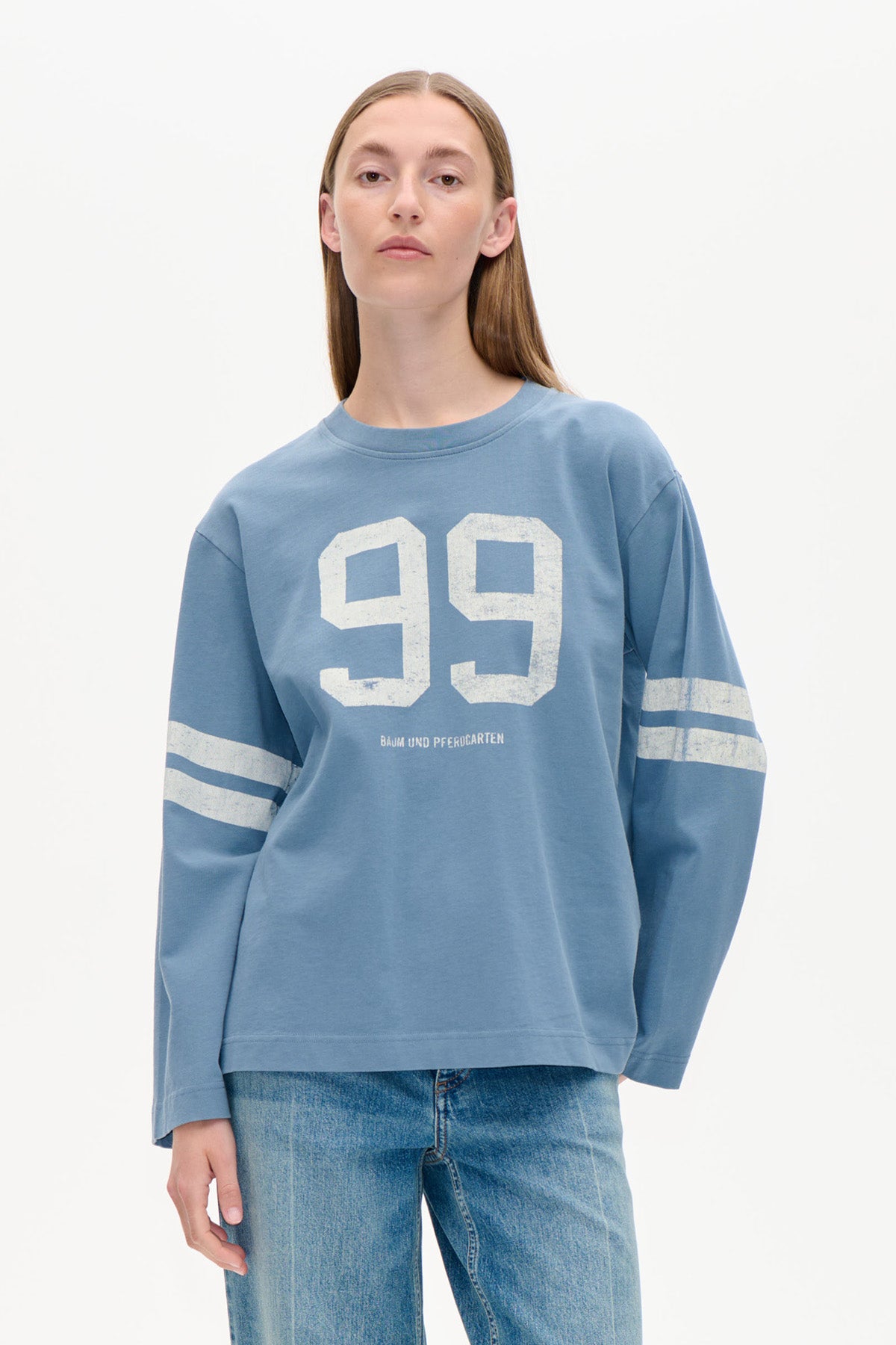 Modell i en dammblå långärmad sweatshirt med vitt '99'-tryck på bröstet och dubbla ränder på ärmarna, stylad med jeans.
