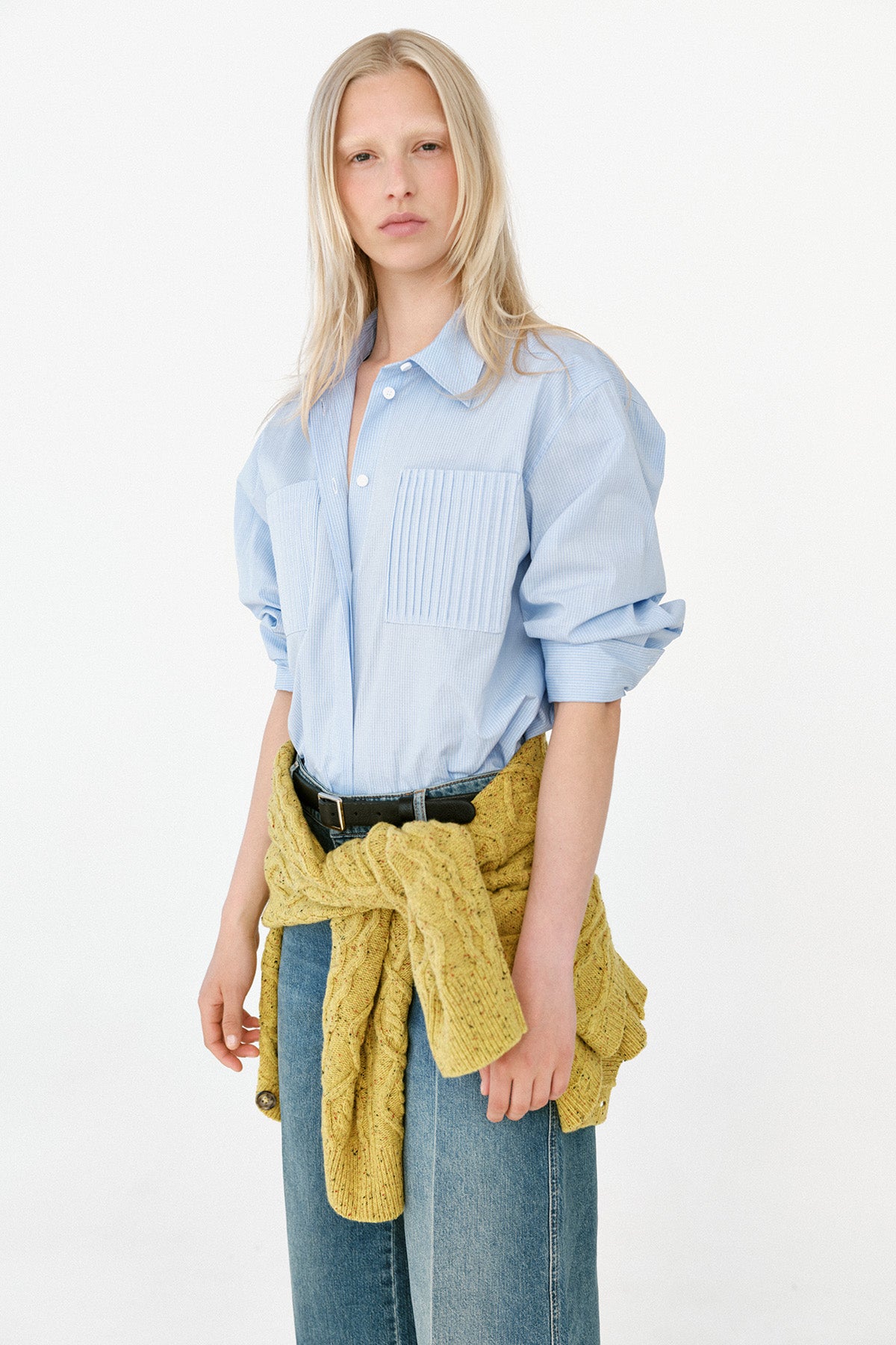 Modellen bär en ljusblå oversized skjorta med plisserad bröstficka, vida blå jeans och en senapsgul stickad tröja knuten i midjan.