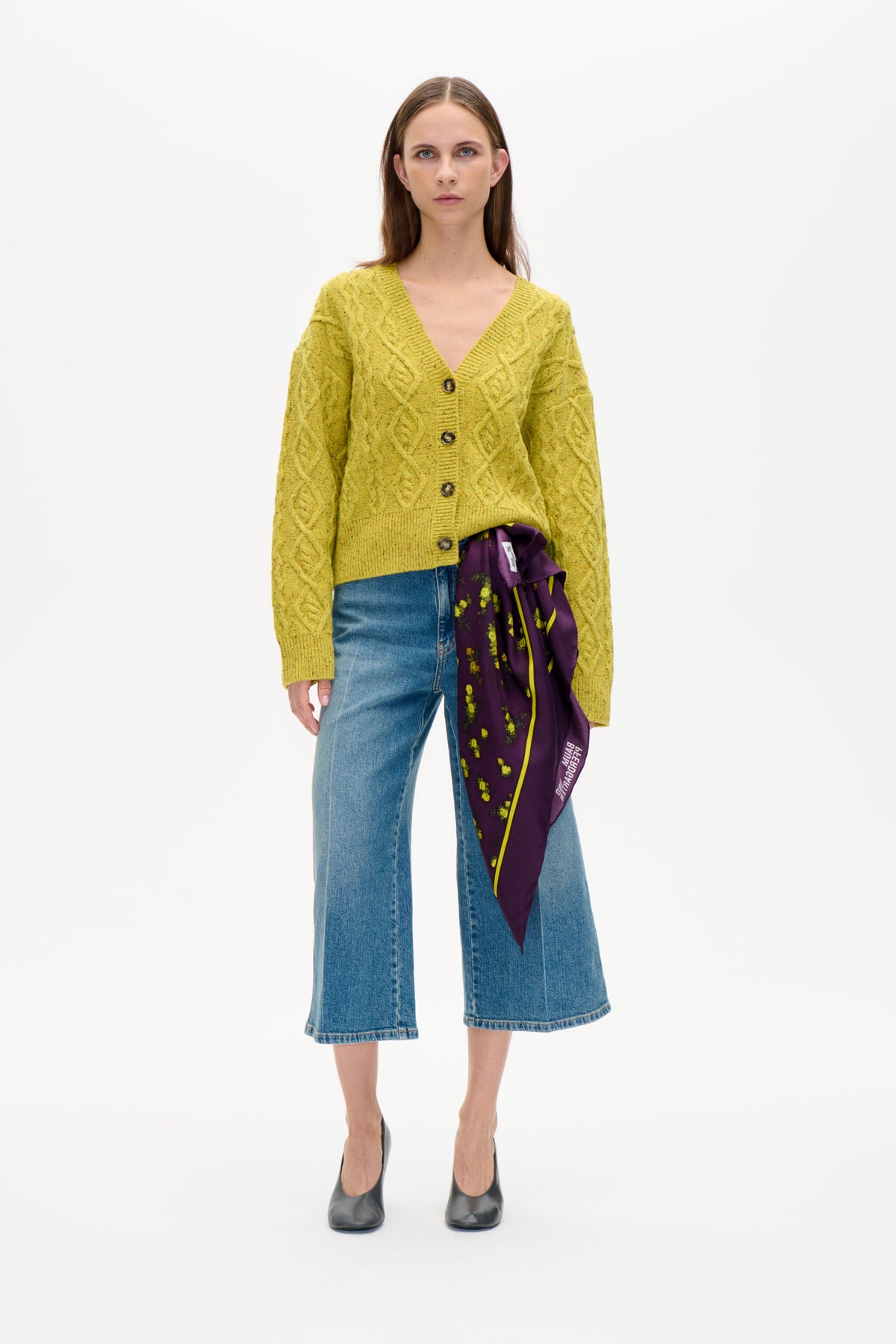 Framifrån: modell i gulgrön flätstickad kofta med knappar, blå vida jeans och lila blommönstrad sjal.