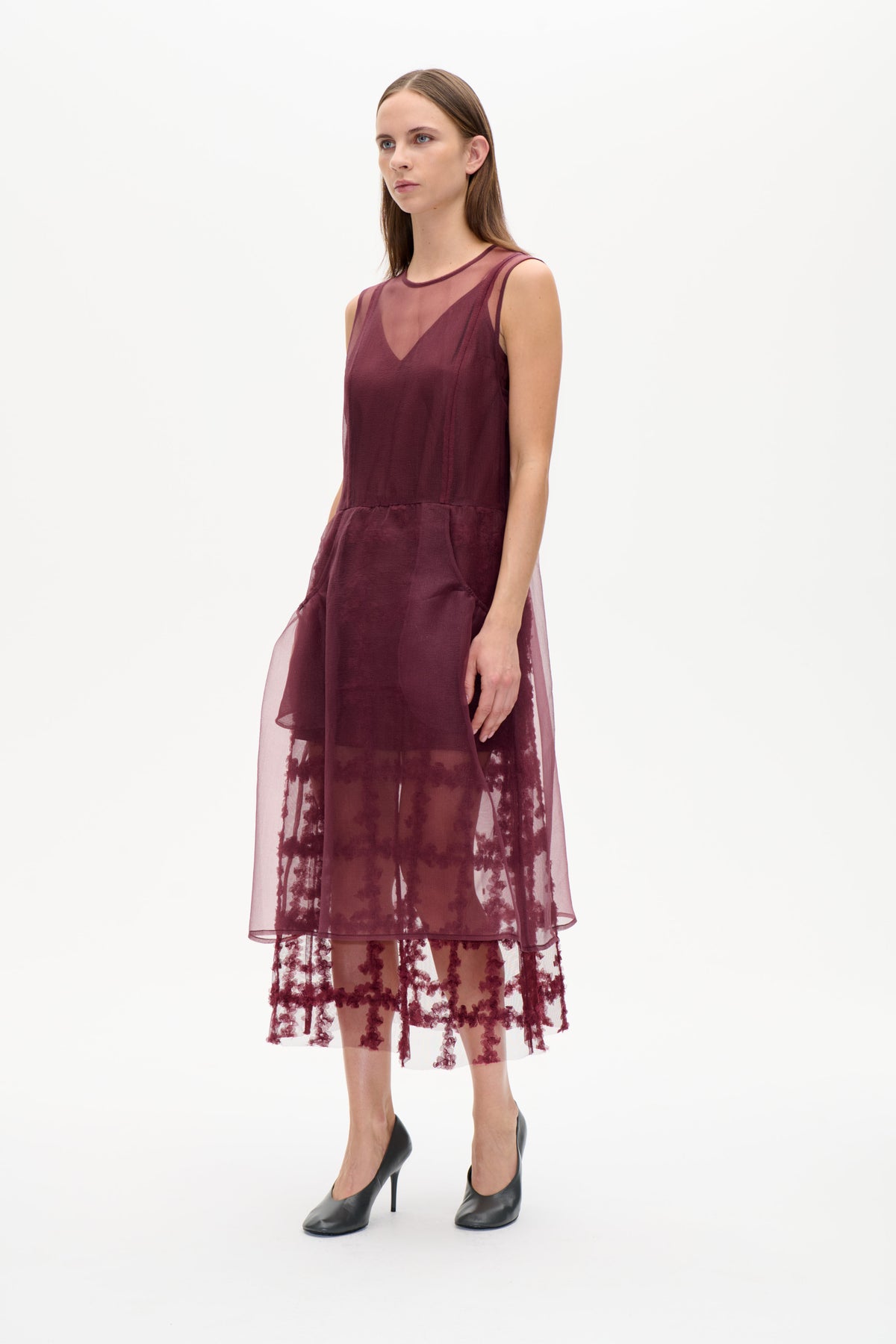 Modell i en plommonfärgad ärmlös klänning med transparent överlager och mörkt broderi med blommotiv, sedd framifrån mot vit bakgrund.