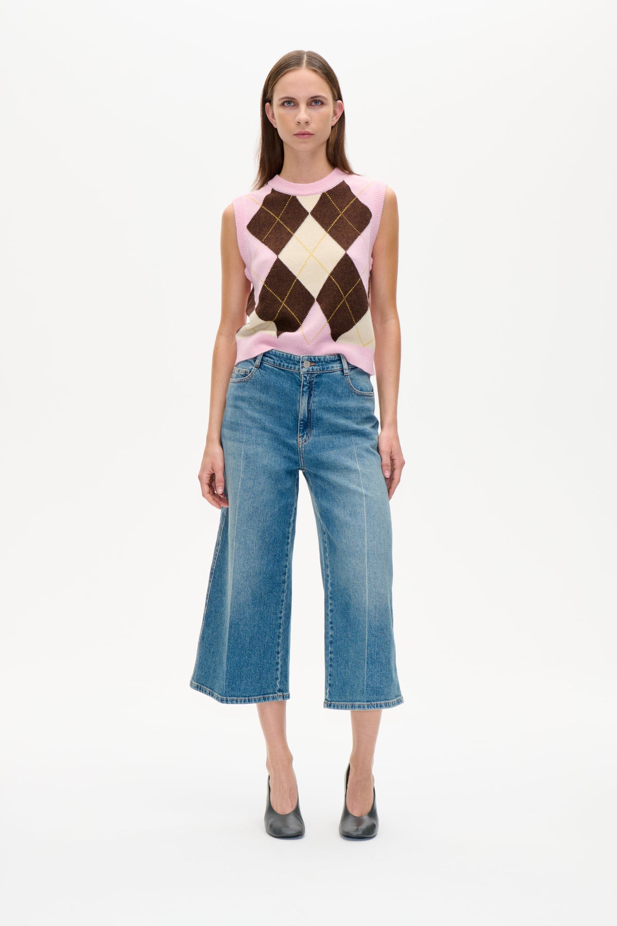Modell som bär en rosa ärmlös stickad väst med bruna och krämfärgade romber, tillsammans med blå vida jeans.