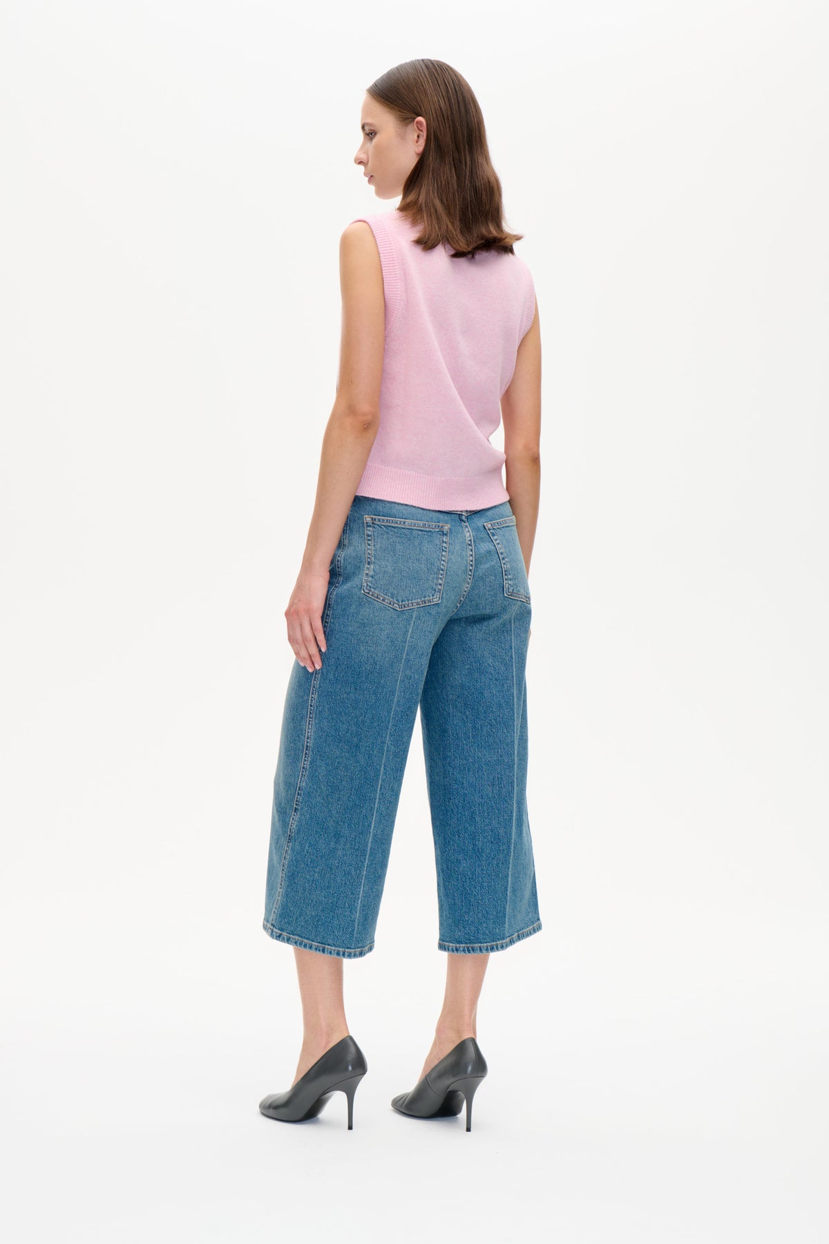 Trekvarts vy bakifrån av en modell som bär en ljusrosa ärmlös stickad väst med ribbad kant och blå vida korta jeans.