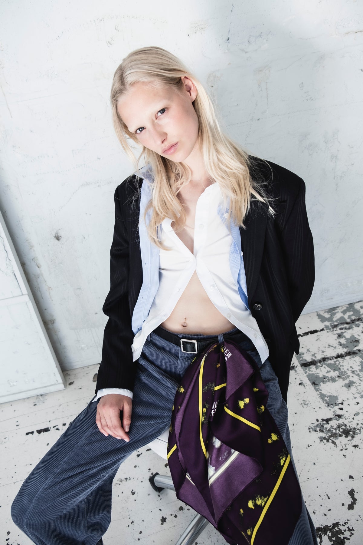 Kampanjporträtt av en modell i en svart blazer, en vit skjorta med krage och en ljusblå smårandig väst, med en lila blommig sjal över mörkblå byxor.
