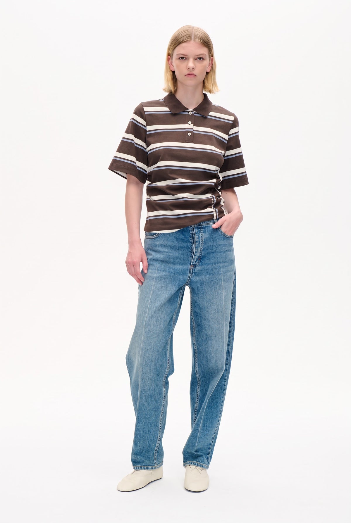 Modell som bär en mörkbrun kortärmad polo med krämfärgade och ljusblå horisontella ränder, kombinerad med ljusblå jeans.