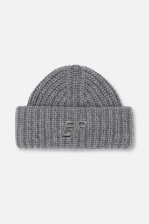 Grå tjockstickad mössa med uppvikt mudd och broderat monogram, sedd framifrån.