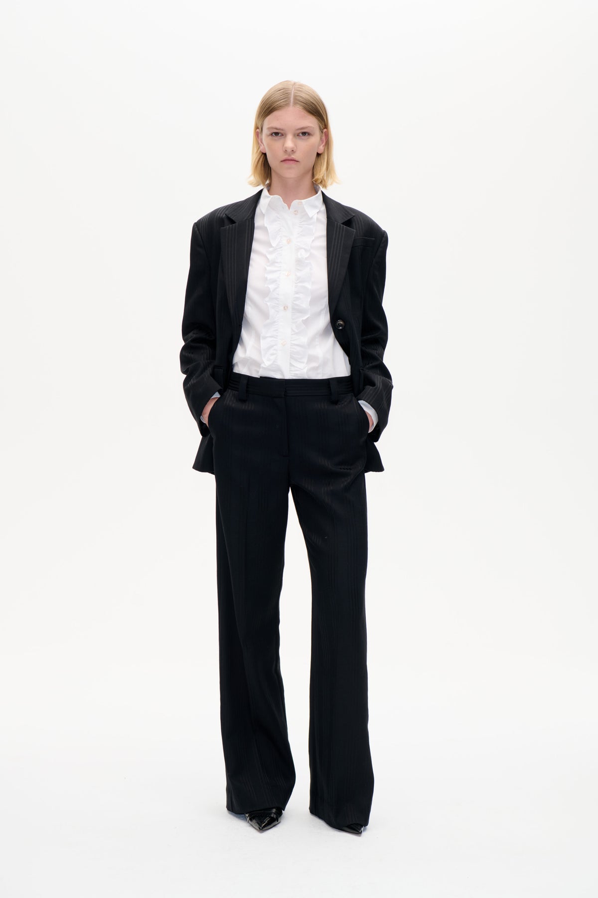 Framåtvänd modell i svart kritstrecksrandig blazer och vida byxor över en vit skjorta med volang, mot en enkel vit bakgrund.