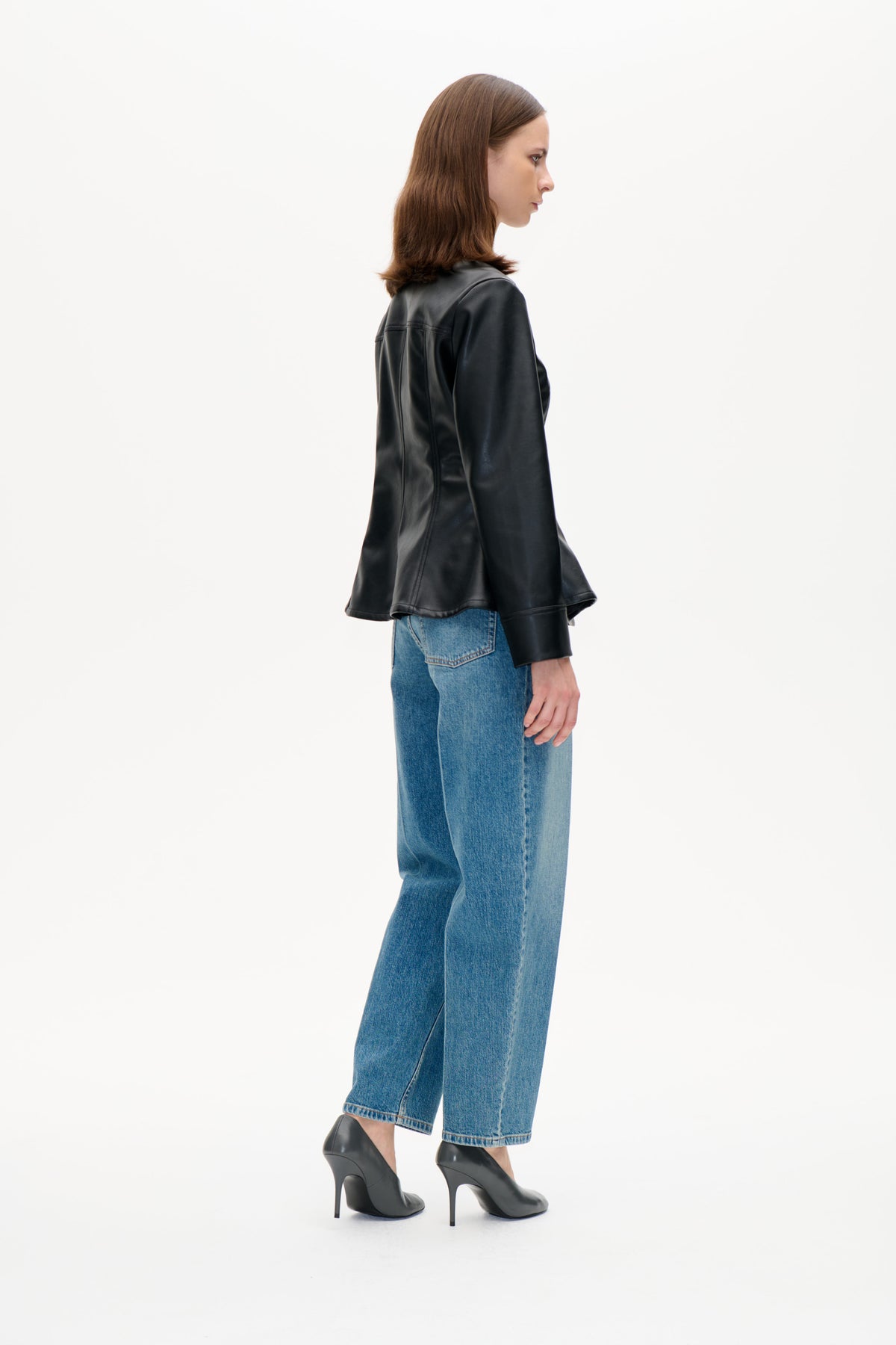Modell i sidoprofil som bär en blank svart jacka med en brun texturerad krage, blå jeans och svarta klackar.