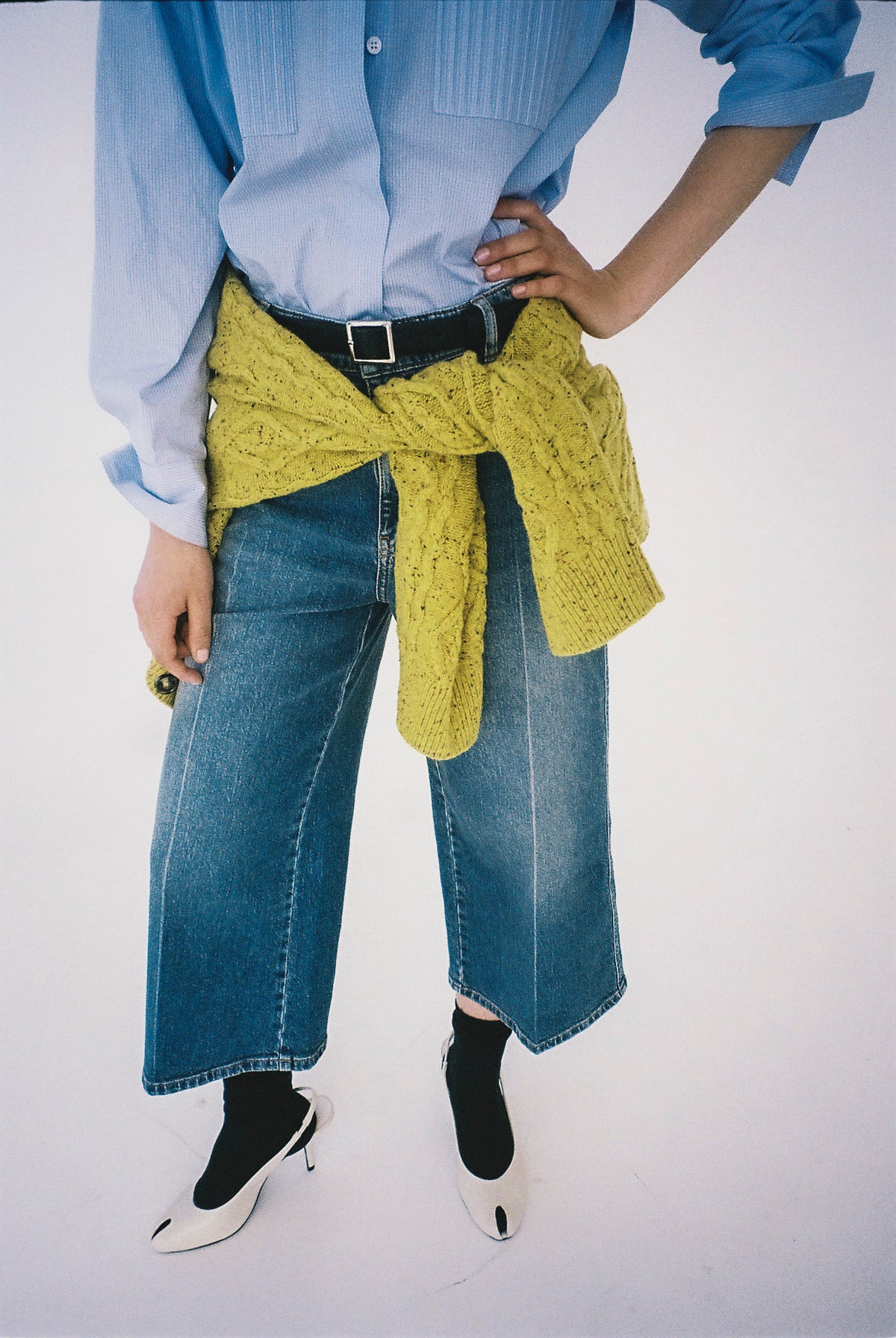 Beskuren kampanjbild av en modell i ljusblå skjorta, vida blå jeans och en senapsgul stickad tröja knuten i midjan.