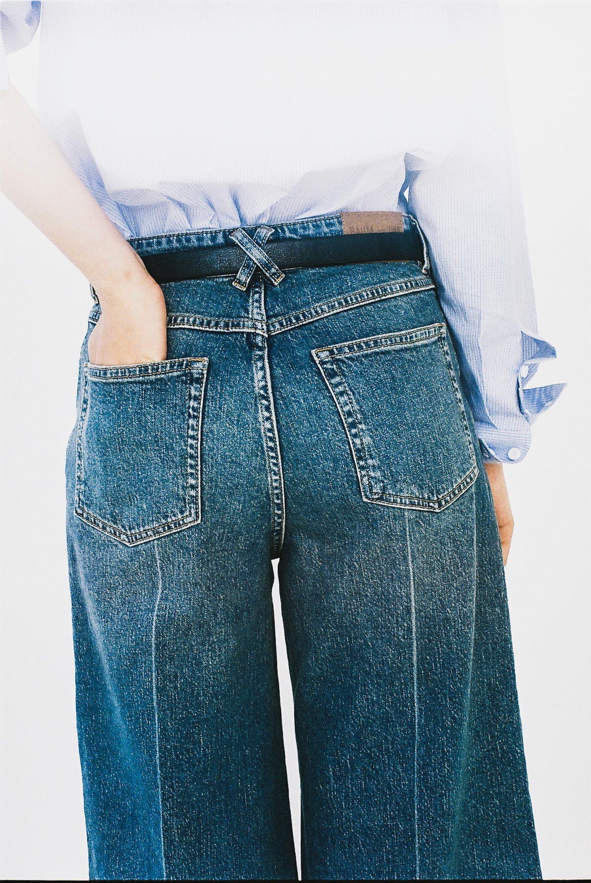 Närbild bakifrån på blå vida jeans med två påsydda bakfickor och en ljusblå randig skjorta instoppad i midjan.