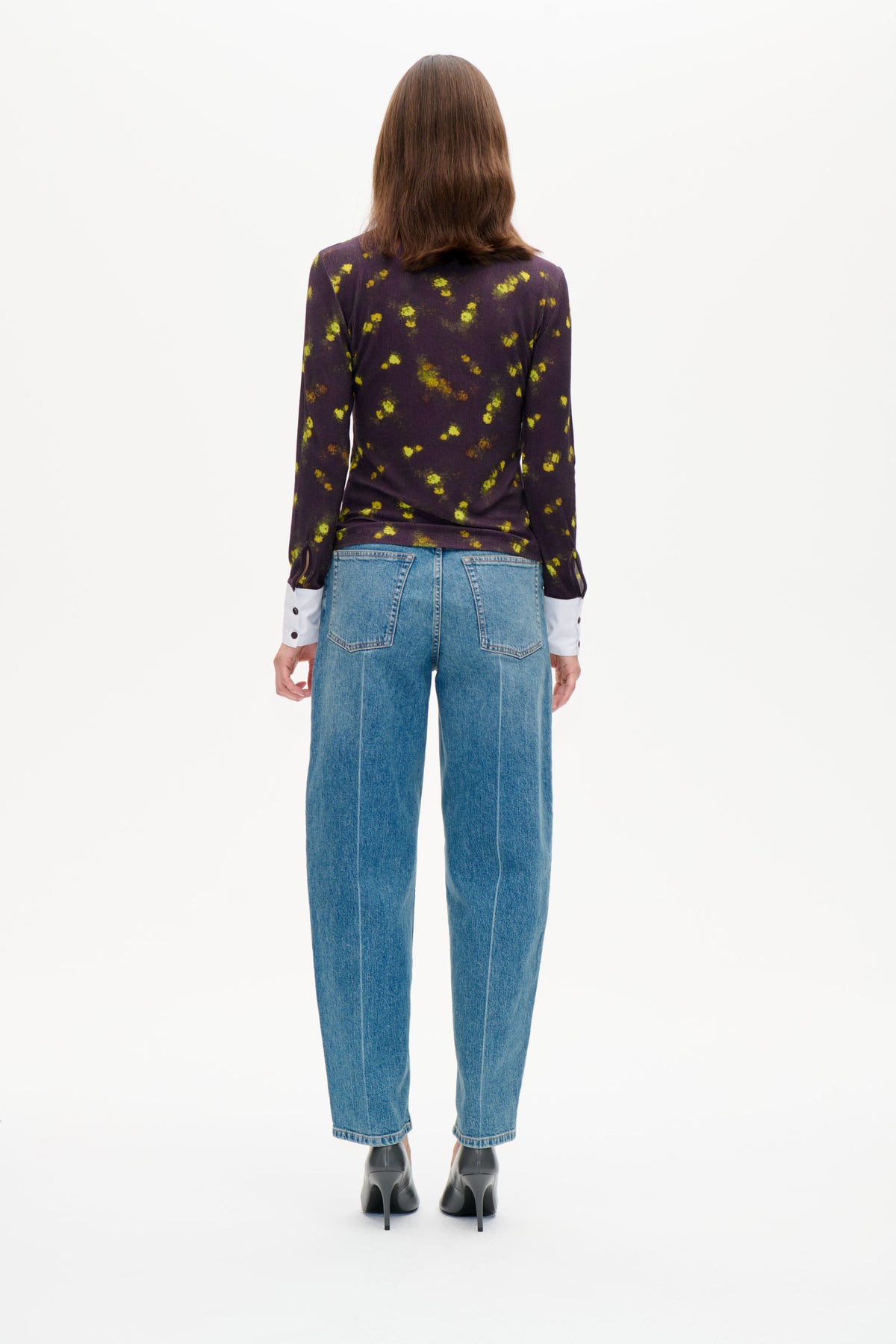 Bakifrån: modell i långärmad mörklila topp med spridda gulgröna blommor, med vita manschetter som syns och blå jeans.