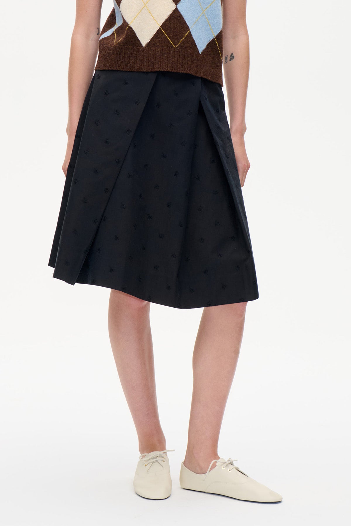 Närbild på en svart knälång kjol med subtila ton-i-tonmönster och överlappande plisserade framstycken, stylad med en stickad väst i rombmönster.