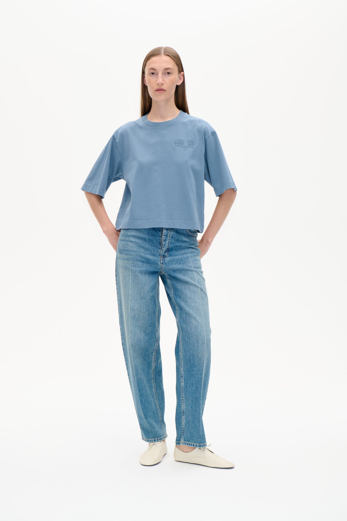 Framifrån en modell i oversized blå t-shirt med rund hals, armbågslånga ärmar och ton-i-ton bröstlogo, matchad med jeans.