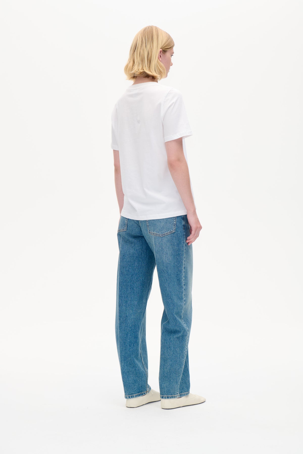Bild bakifrån av en modell som bär en slät vit kortärmad t-shirt, blå jeans och vita skor mot en vit bakgrund.