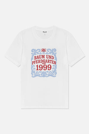 Framifrån: en vit kortärmad t-shirt med en blå dekorativ ram, en röd blomdetalj och texten 'Baum und Pferdgarten 1999'.