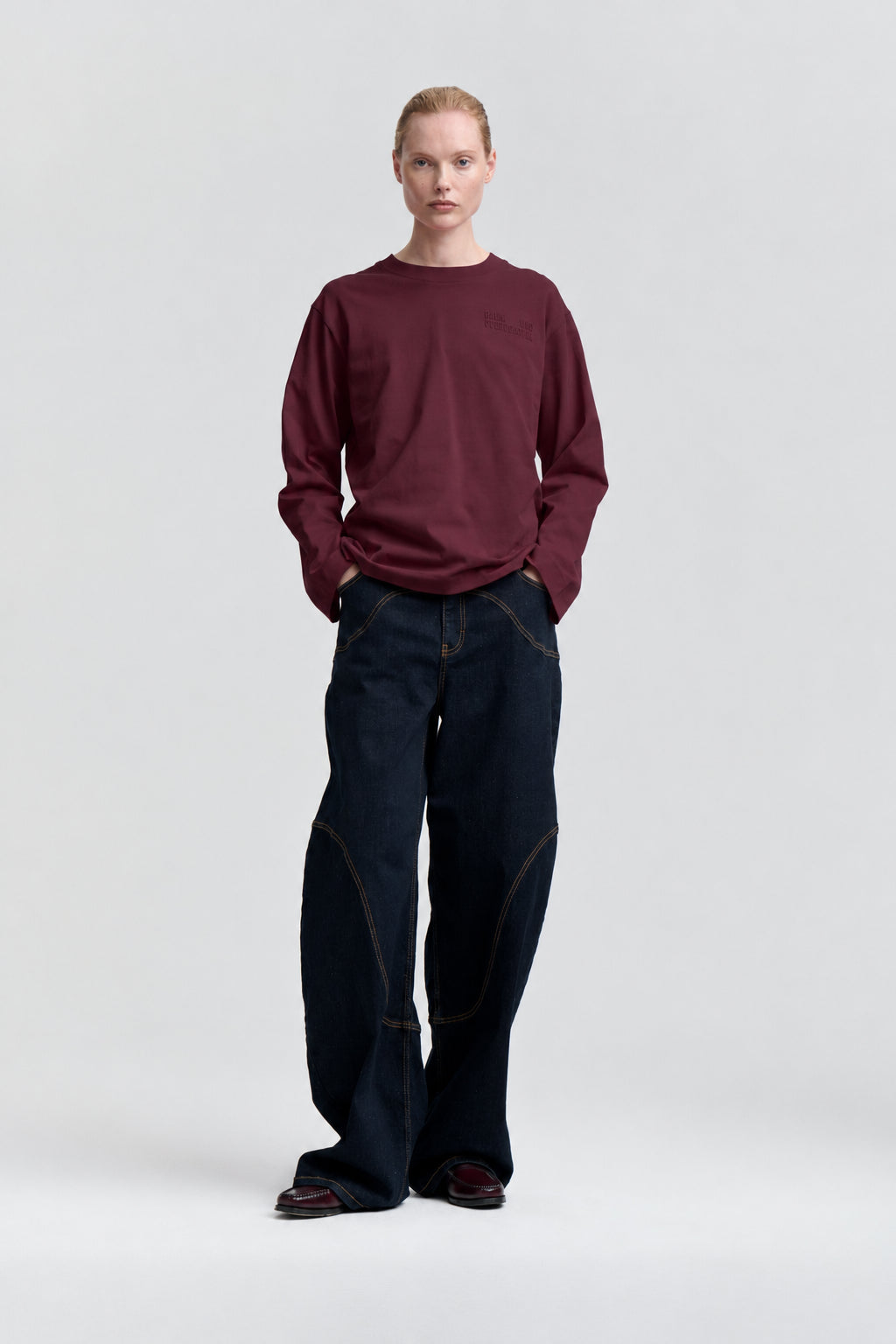 Framifrån: modell i en vinröd långärmad sweatshirt och vida mörk indigoblå jeans med kontrastsömmar.