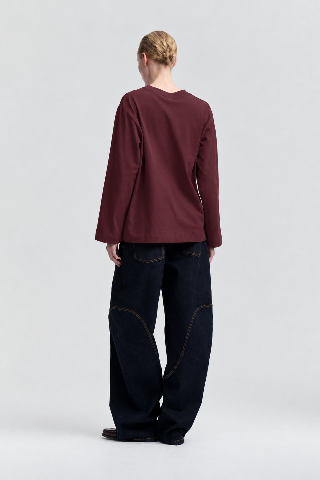 Modell sedd bakifrån i en enfärgad vinröd långärmad topp med rund hals och mörka vida jeans.