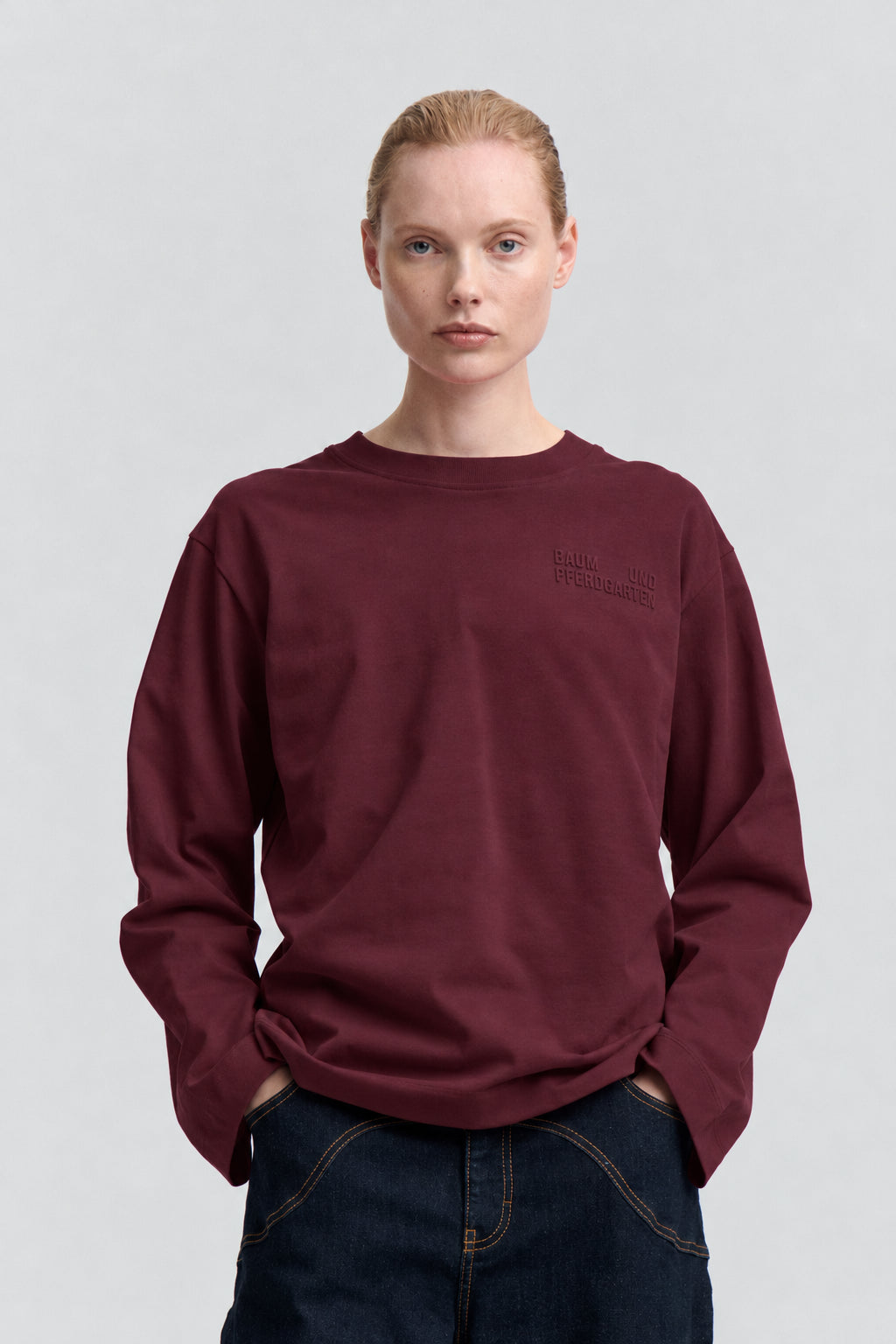 Framifrån: en vinröd långärmad sweatshirt med rund hals och subtil ton-i-ton brösttext, kombinerad med mörka jeans.