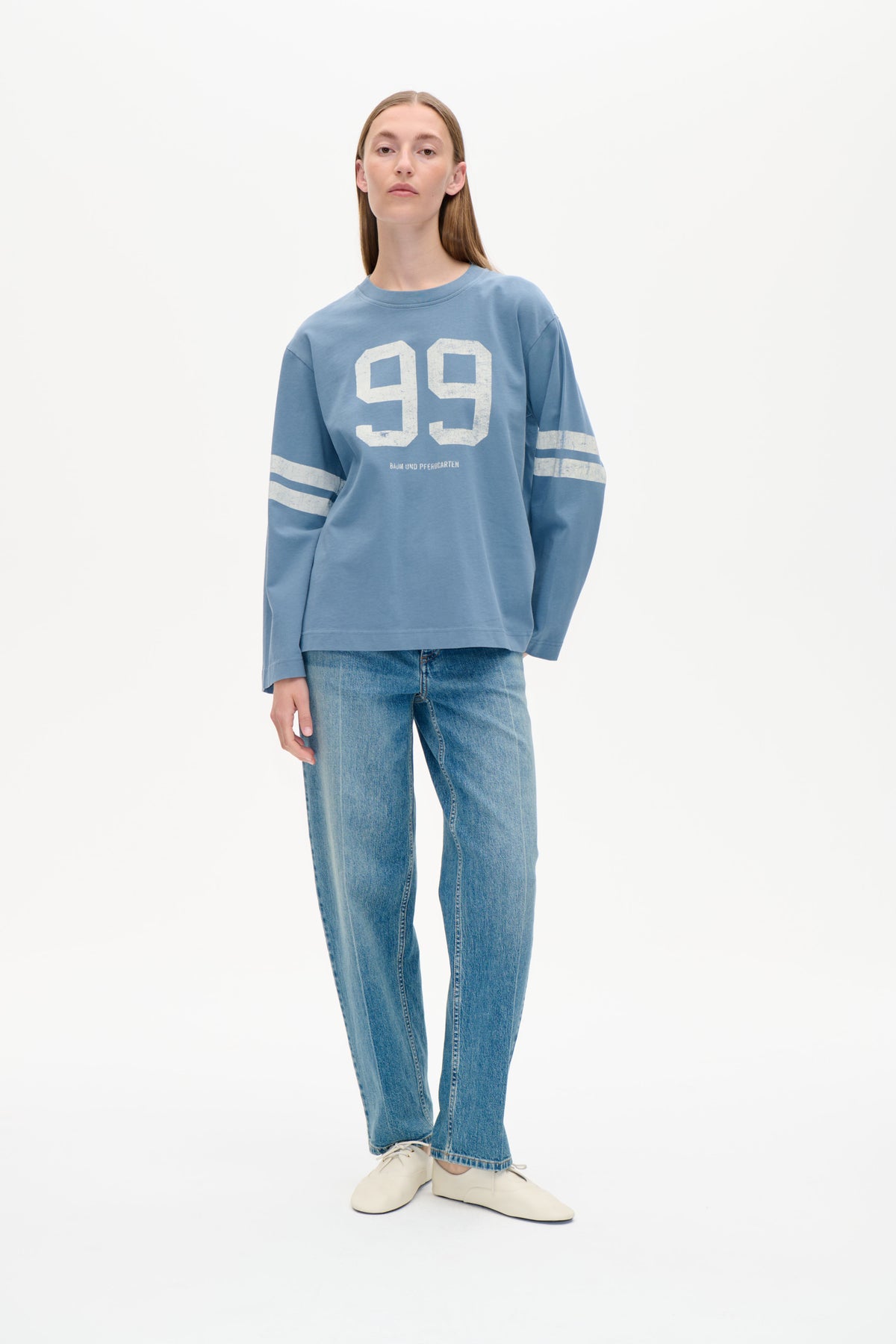 Framifrån: modell i en vid blå sweatshirt med stort slitet vitt 99-tryck och randiga ärmar, matchad med jeans.
