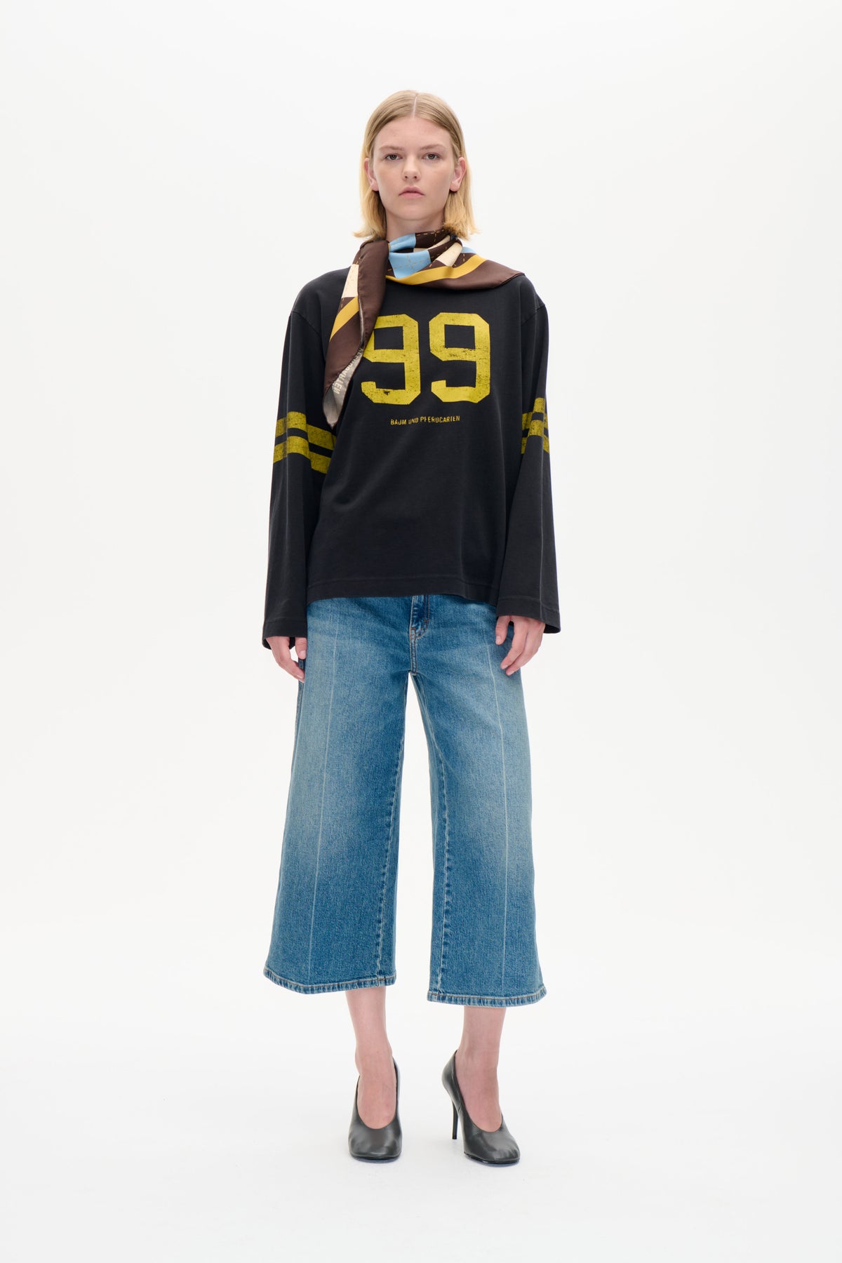 Modellen bär en mörkgrå långärmad sweatshirt med ett stort gult 99-tryck, vida blå jeans och en flerfärgad geometrisk sjal.