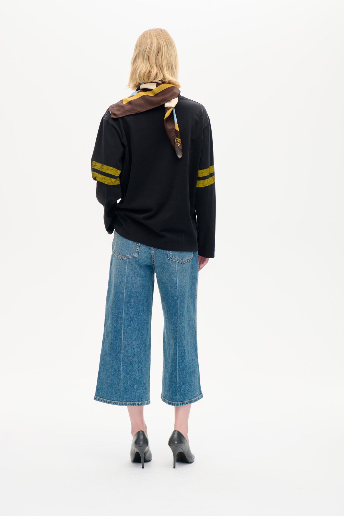 Bakifrån av en modell som bär en svart långärmad topp med senapsgula ränder på ärmarna, en flerfärgad geometrisk sjal och vida blå jeans.