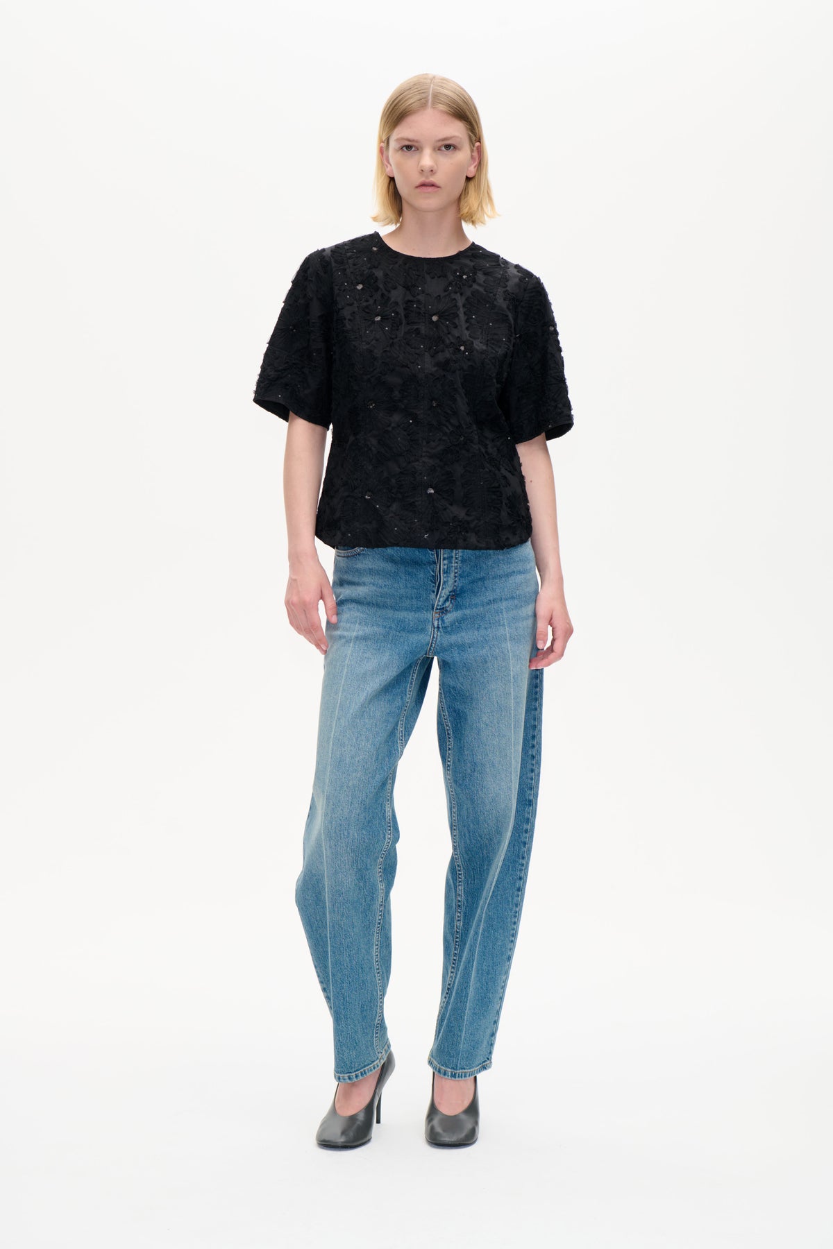Modell sedd framifrån i en svart kortärmad topp med upphöjda blomdetaljer och små prydnader, matchad med blå jeans.