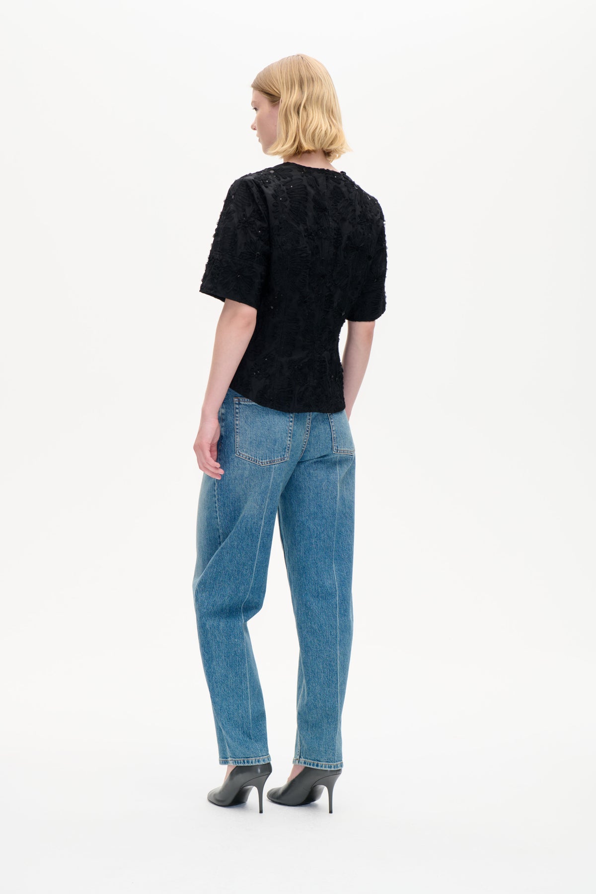 Modell sedd bakifrån i en svart kortärmad topp med texturerade blomdetaljer och blå jeans.