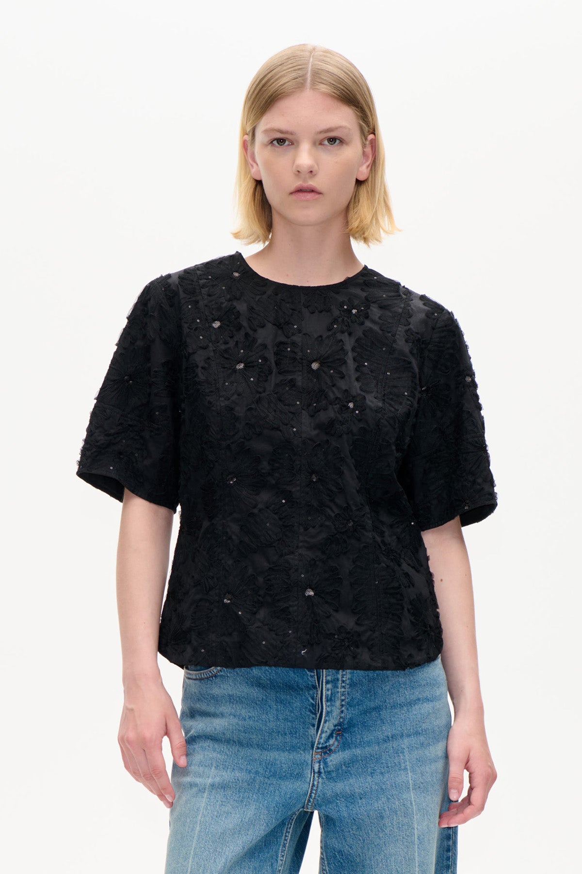 Framifrån en svart kortärmad topp med upphöjda blomdetaljer och små dekorativa inslag, matchad med blå jeans.