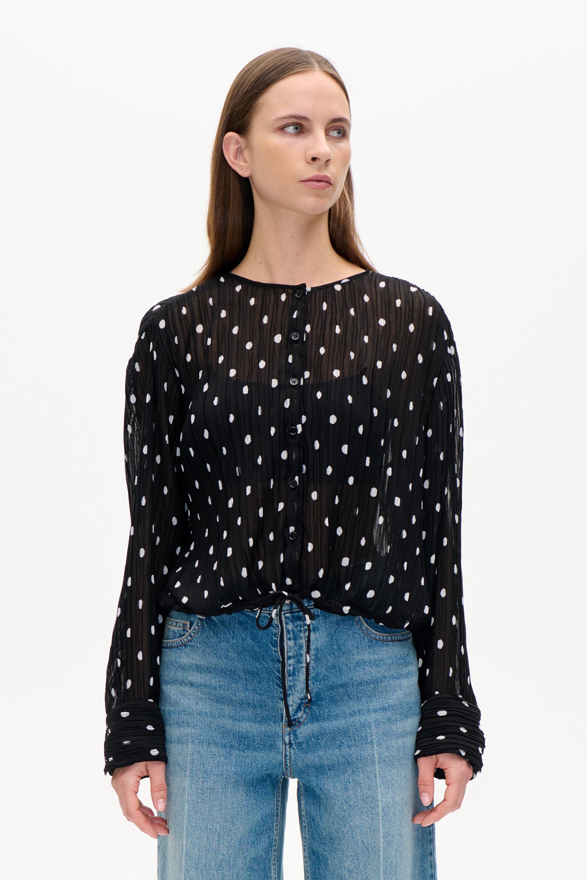Framsida av en svart transparent krinklad blus med vita oregelbundna prickar, knäppning framtill och långa ärmar, bärs med blå jeans.