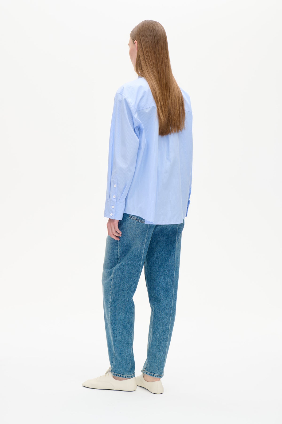 Bakifrån: modell som bär en ljusblå, finrandig, långärmad skjorta med knappar och blå jeans.