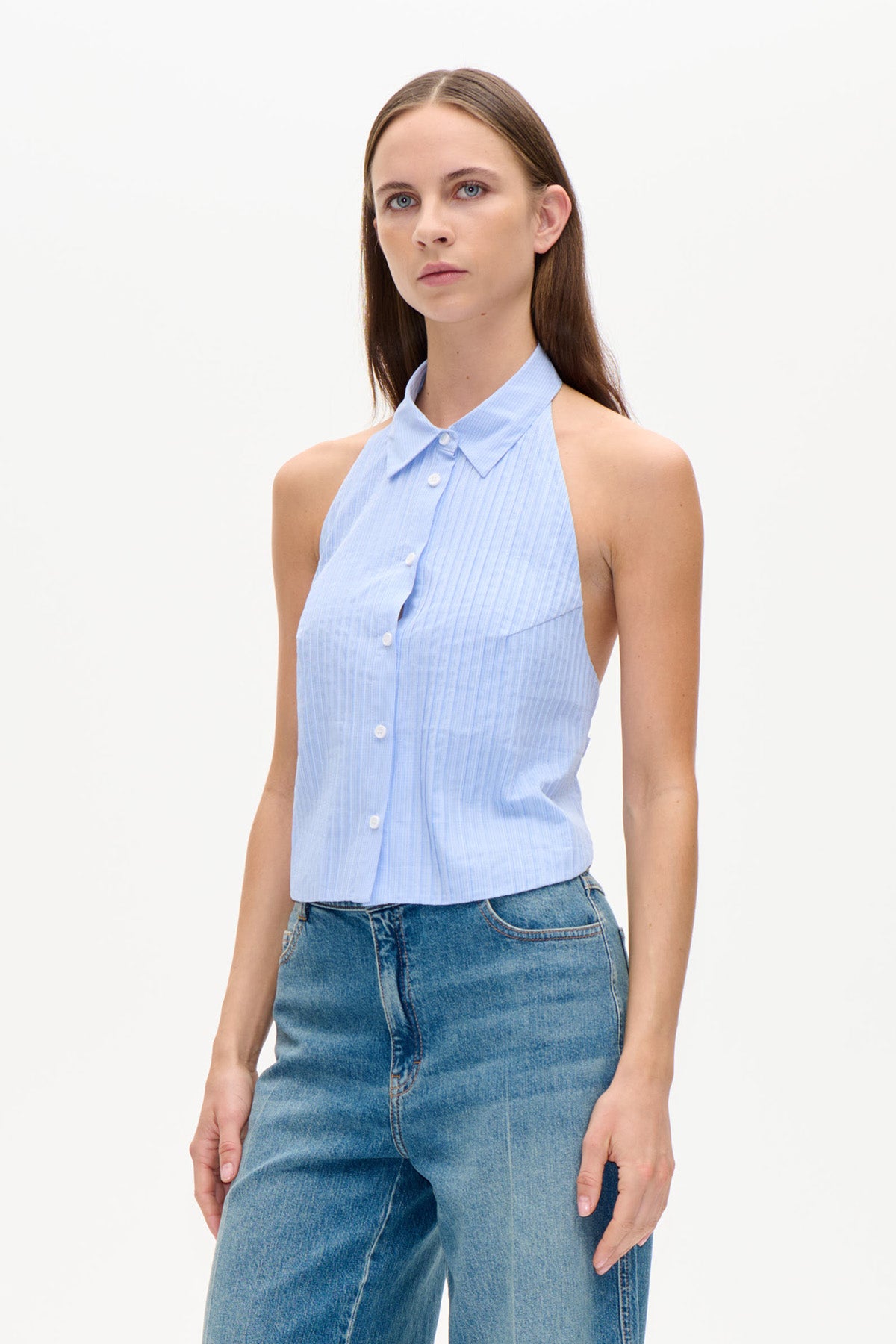 Modellen bär en ljusblå, fint randig ärmlös topp med krage, knäppning fram och halterneckringning, tillsammans med blå jeans.