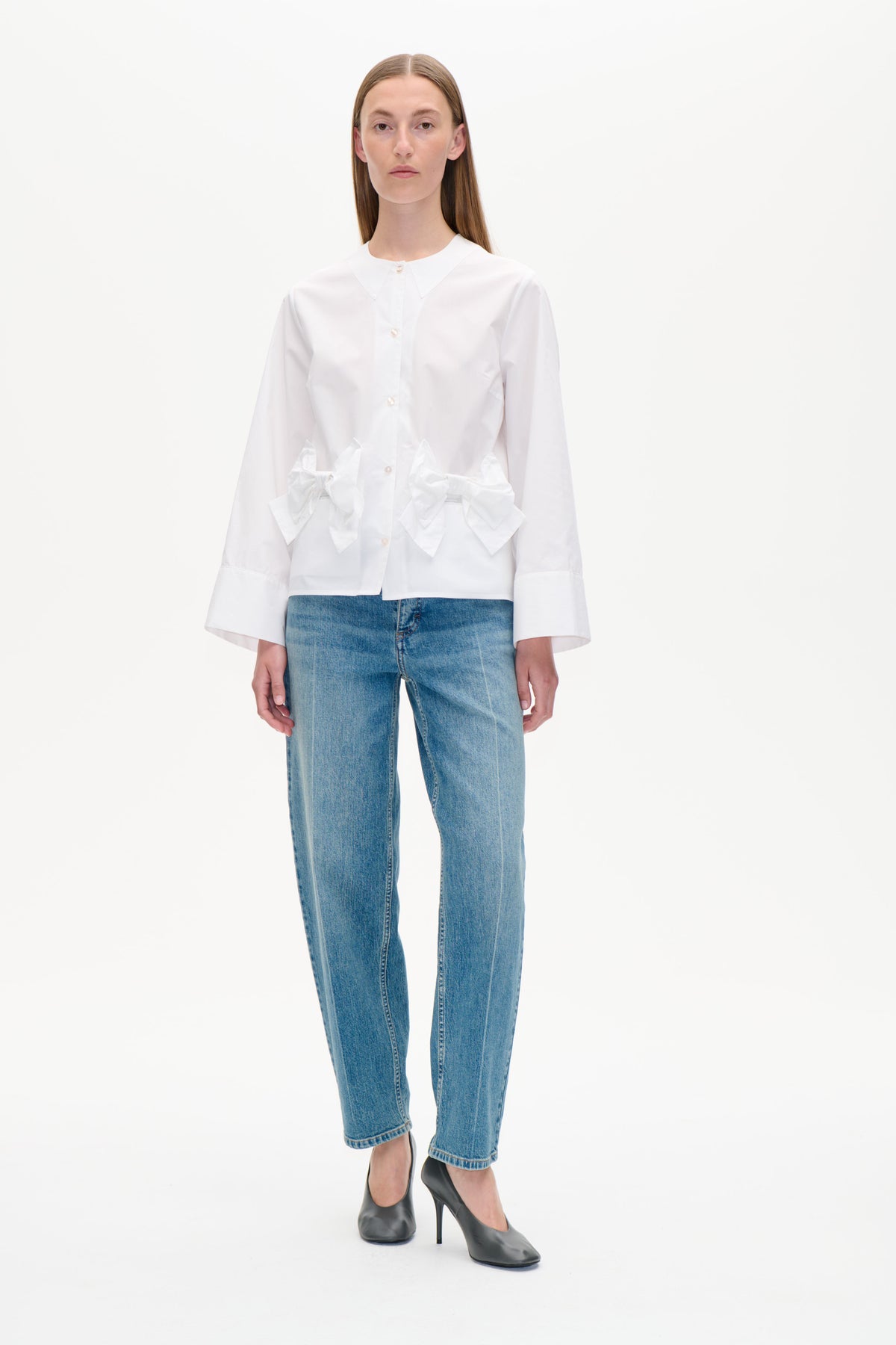 Framifrån: en vit skjorta med knappslå, oversized spetsig krage och skulpturala rosettdetaljer i midjan, kombinerad med blå jeans.