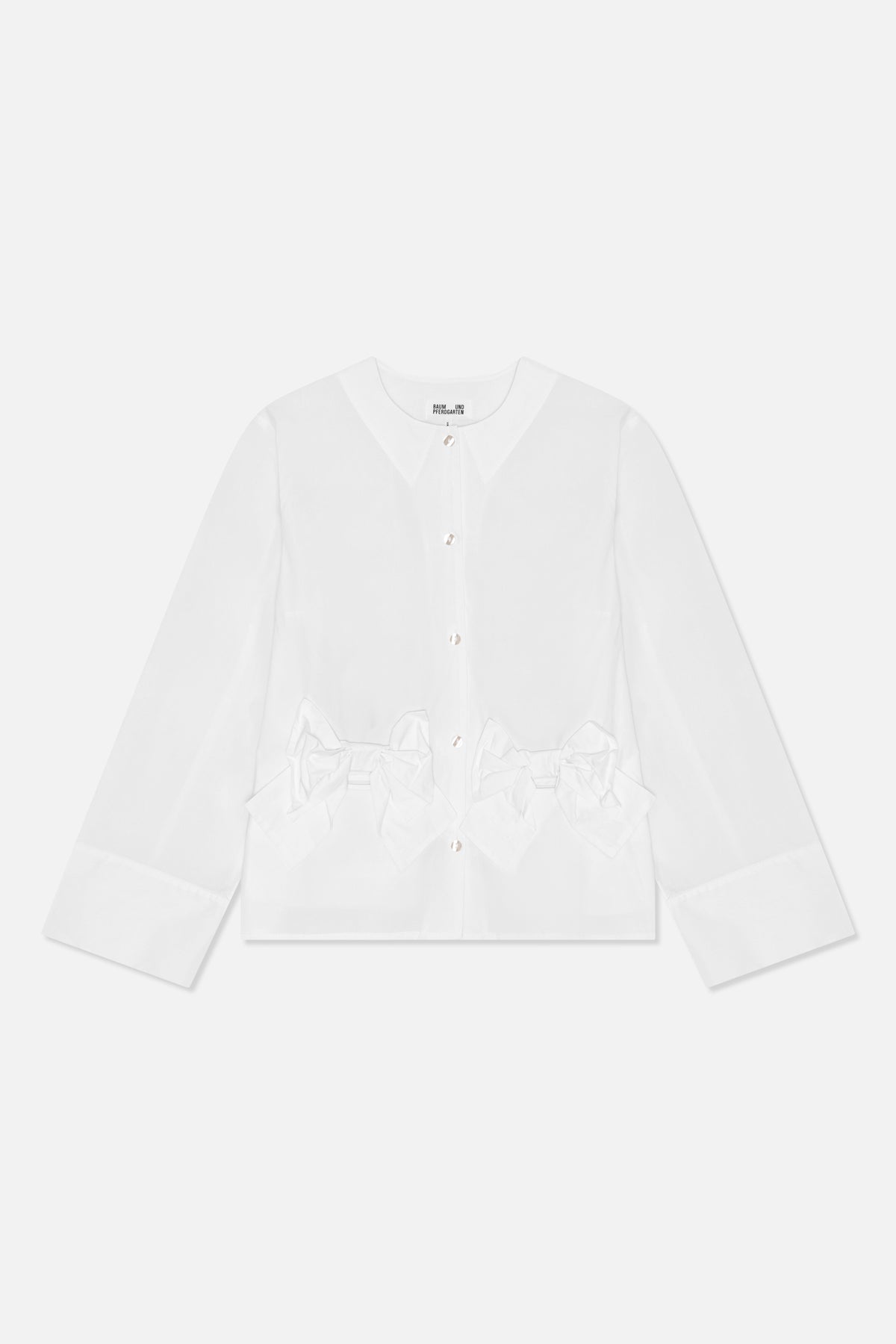 Framifrån en vit långärmad blus med rund hals, blanka knappar, vida manschetter och oversized rosettdetaljer i midjan.