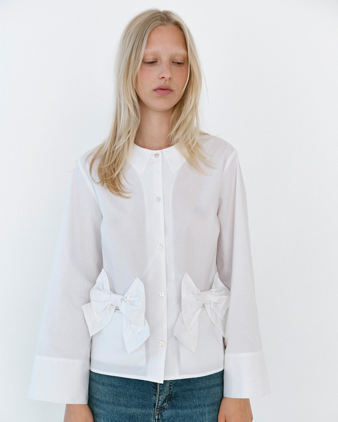 Modellen bär en vit blus med knäppning fram, spetsig krage, oversized rosettdetaljer i midjan och vida manschetter, matchad med blå jeans.