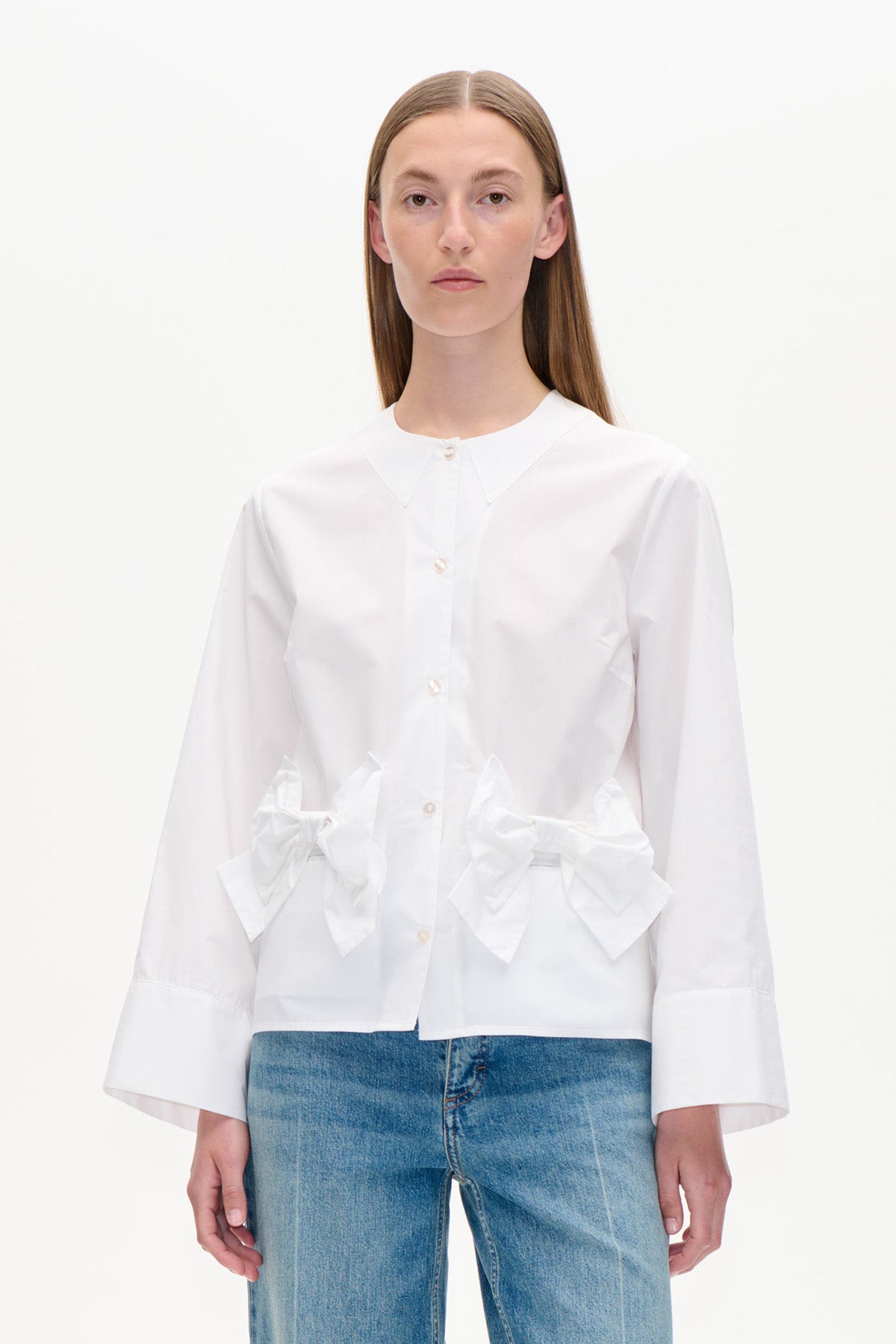 Framifrån en vit knäppskjorta med spetsig krage, oversized manschetter och stora rosetter i midjan, matchad med blå jeans.