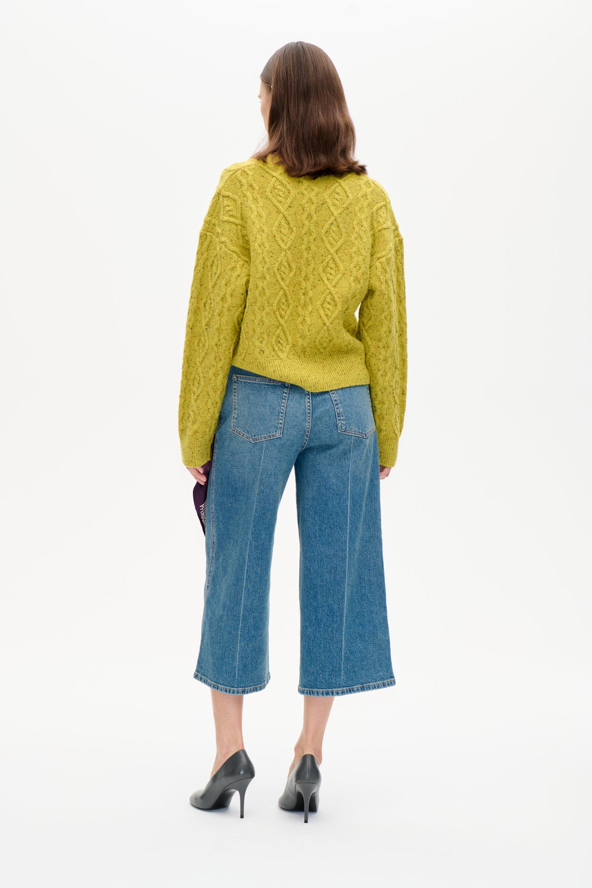 Bakifrån: modell i en gulgrön melerad flätstickad tröja och blå croppade vida jeans.