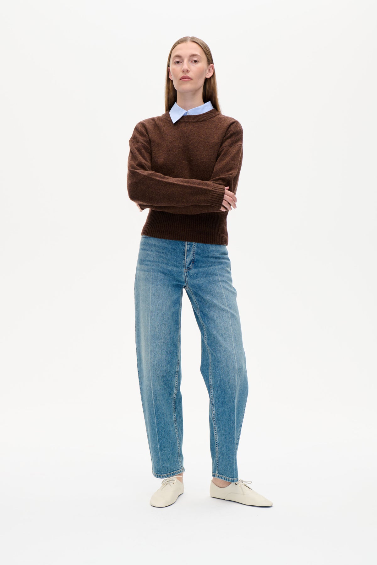 Framifrån syns en modell i en brun rundhalsad tröja över en ljusblå randig skjorta med krage, blå jeans och krämfärgade snörskor.