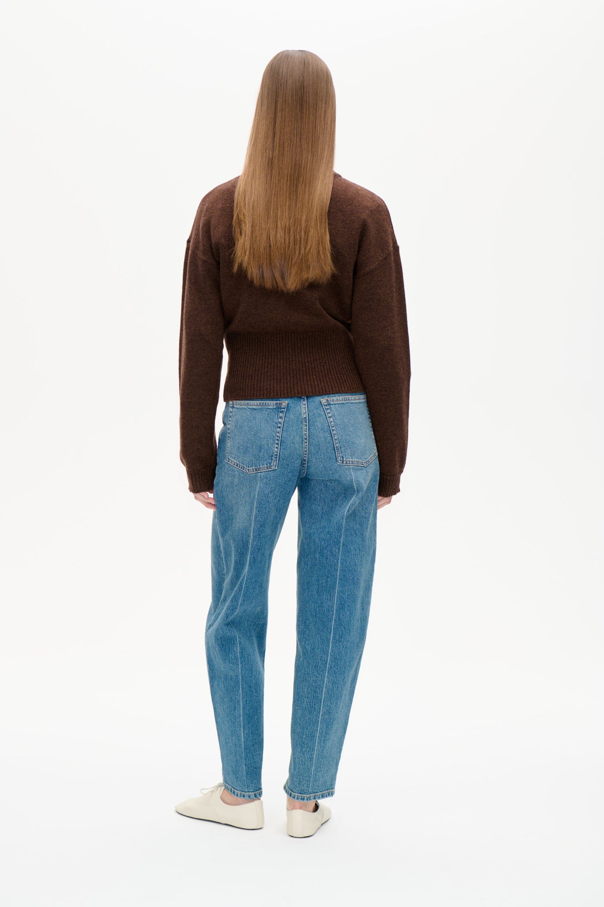 Modell fotograferad bakifrån i mörkbrun långärmad stickad tröja med ribbad nederkant och blå jeans.