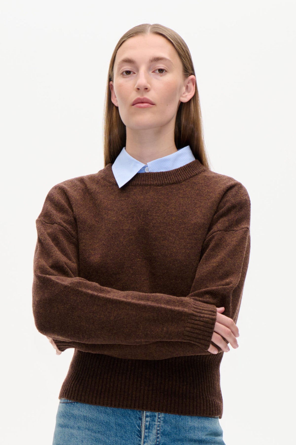 Framifrån: en modell i en brun stickad crewnecktröja över en ljusblå skjorta och blå jeans.