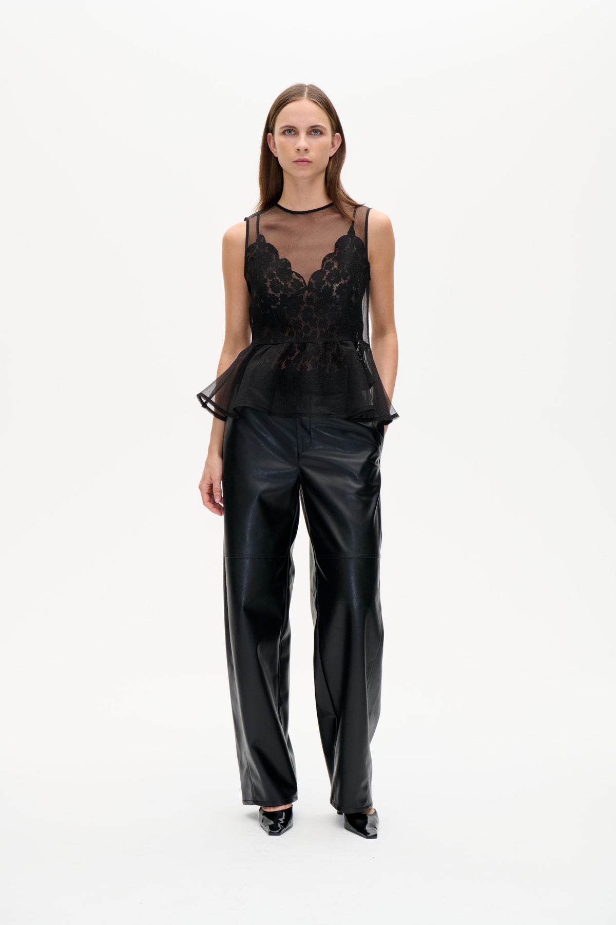 Modellen bär en svart transparent ärmlös topp med blommönstrade spetspaneler och utsvängd peplum, kombinerad med glansiga vida byxor.
