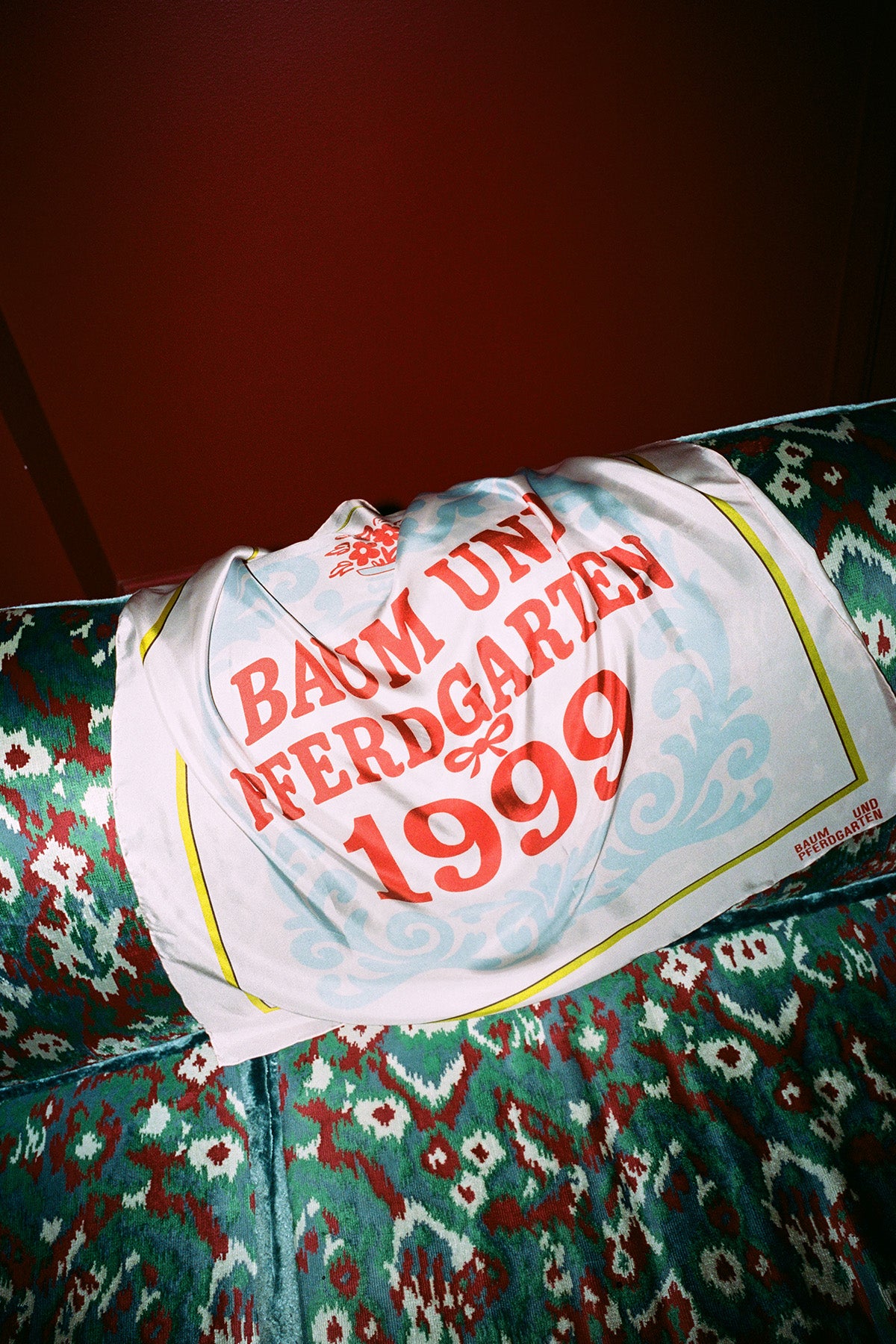 Tryckt kvadratisk sjal draperad över en mönstrad stol, med blekblå snirkliga mönster, gul kant och röd text "Baum und Pferdgarten 1999".